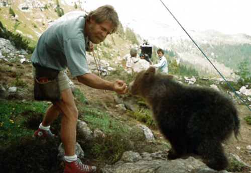 A man in shorts feeds a bear in a mountainous landscape, with others observing in the background.